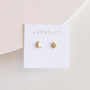 A white card labeled JaxKelly displays the Sun & Moon earrings—gold CZ studs shaped like a crescent moon with a gem and a radiant sun—on a light, textured surface.