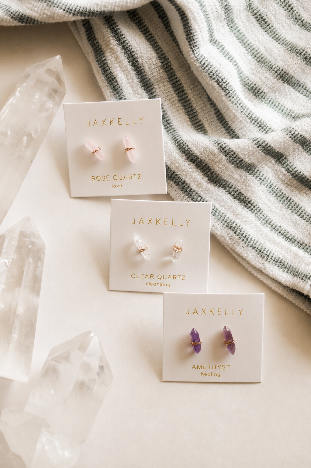 A JaxKelly Clear Quartz Mineral Point for cleansing is displayed on a light surface with crystals and striped fabric in the background.
