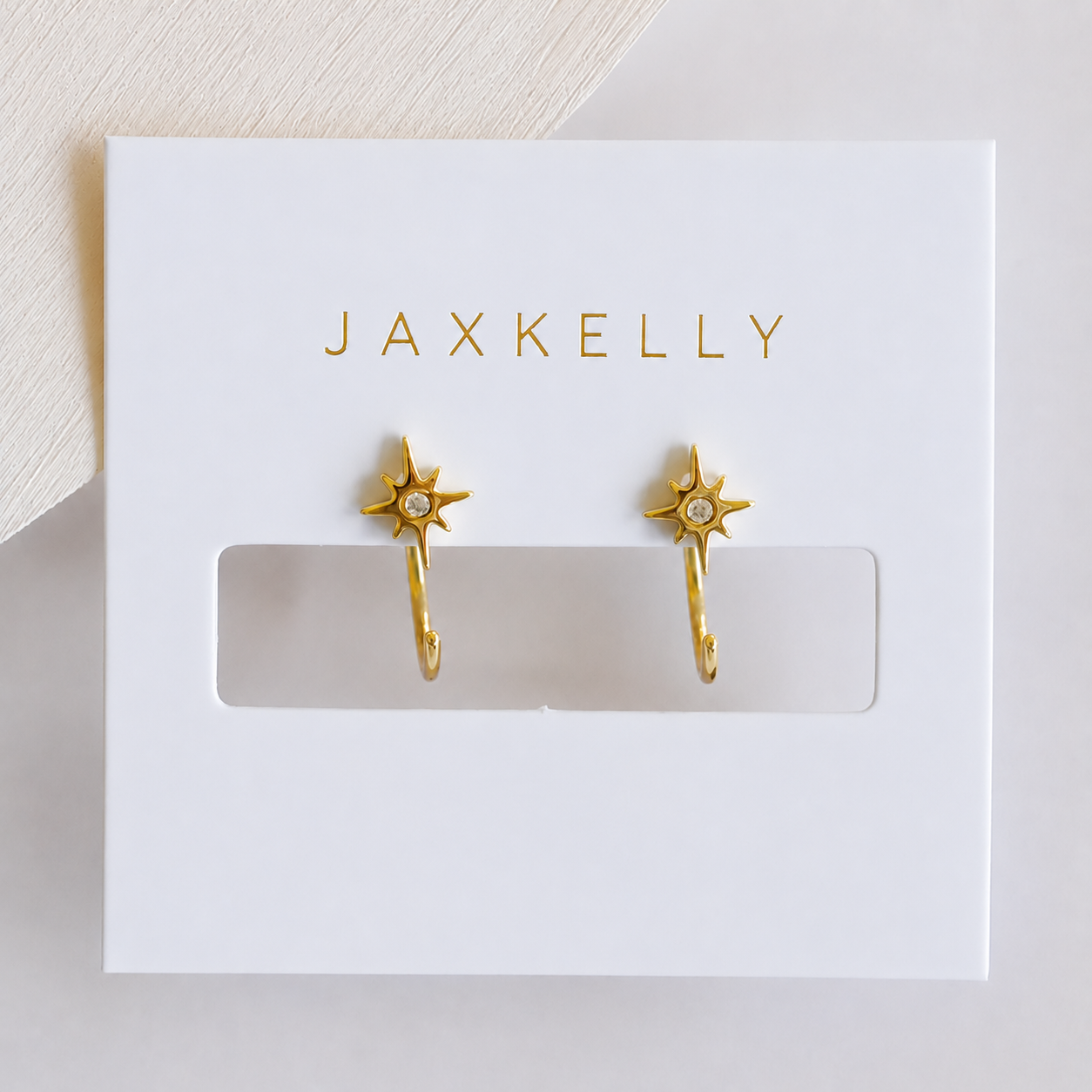 The JaxKelly Star Huggie earrings feature an 18K gold-plated starburst design with a clear center stone, displayed on a white card with "JAXKELLY" in gold letters.