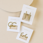 Three pairs of JaxKelly gold hoop earrings, including the Tiny Ball Hoop ideal for ear stacks, are shown on white JaxKelly cards against a beige background. Each card features a unique hypoallergenic design.