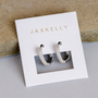 A pair of Pavé Silver Hoops by JaxKelly, adorned with small crystals, shown on a white card with gold JaxKelly lettering, resting on a light tan textured surface.