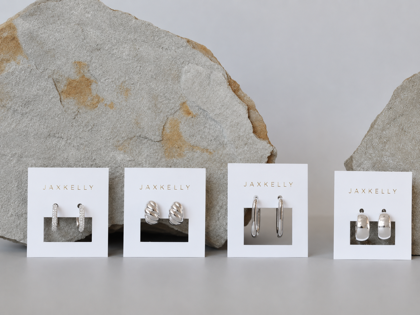 Four pairs of JaxKelly earrings, including Pavé Silver Hoop styles, are displayed on branded cards before two large rocks on a light gray background. The cards feature unique rhodium-plated designs from hoops to detailed studs.