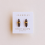 A pair of smoky quartz mineral point earrings by JaxKelly, labeled "Smoky Quartz Mineral Point - Protection," are displayed on a white card with gold and gray lettering.