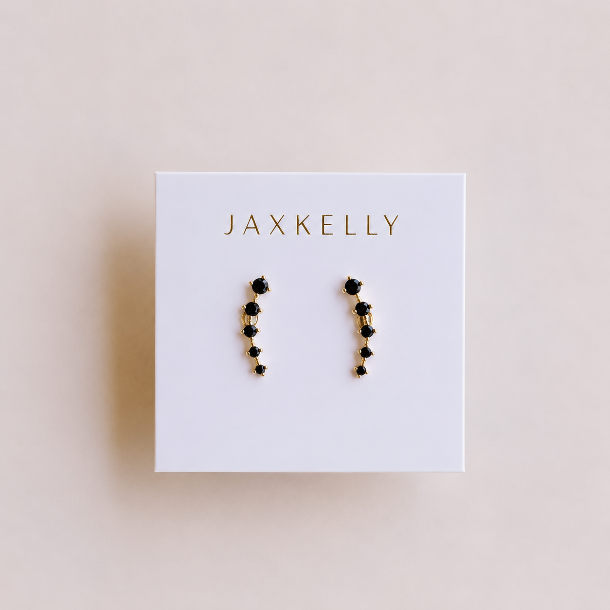 The Black Crawler earrings by JaxKelly feature black stones in a curved line with 18k gold plating, displayed on a white box with JaxKelly in gold lettering against a soft beige background.