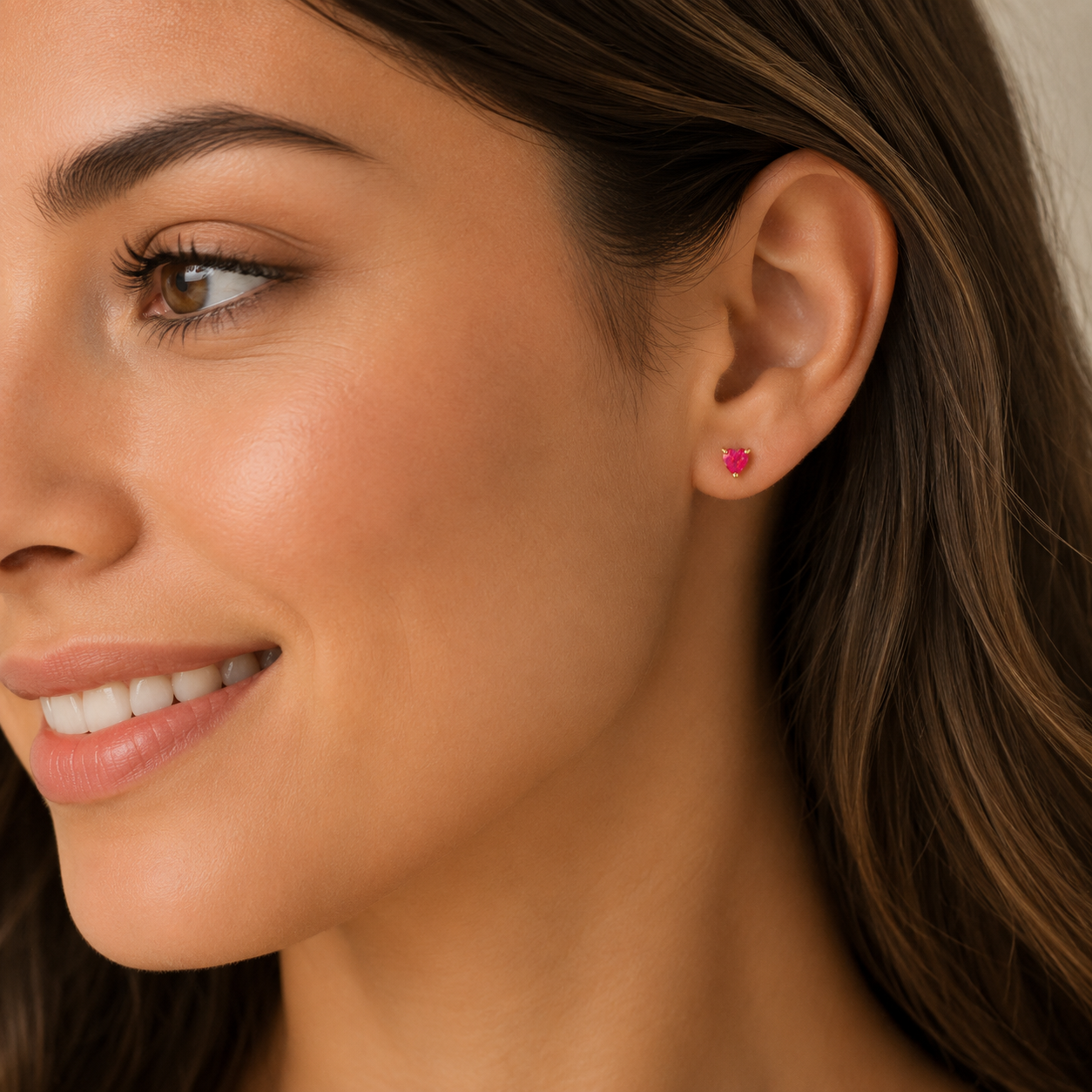 A woman with long brown hair smiles softly in profile, wearing JaxKelly's Puffed Heart - Pink Opal stud earring. The heart-shaped design and 18k gold plating stand out against the neutral, gently blurred background.