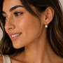 A woman with long brown hair and radiant skin smiles softly, wearing JaxKelly’s Gemstone Mini Drop - Rose Quartz gold plated earrings, her natural makeup enhancing the elegant look as she looks slightly to the side.