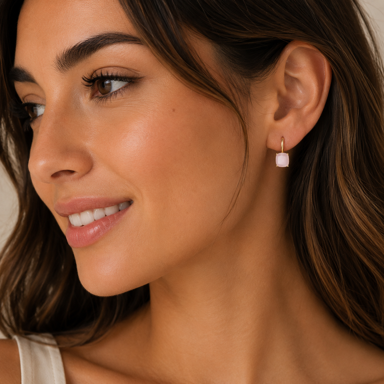 A woman with long brown hair and radiant skin smiles softly, wearing JaxKelly’s Gemstone Mini Drop - Rose Quartz gold plated earrings, her natural makeup enhancing the elegant look as she looks slightly to the side.
