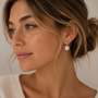 A woman with light brown hair in a loose bun, wearing a cream sweater and JaxKelly’s Gemstone Mini Drop - Mother of Pearl earrings, gazes thoughtfully to the side against a soft beige background.