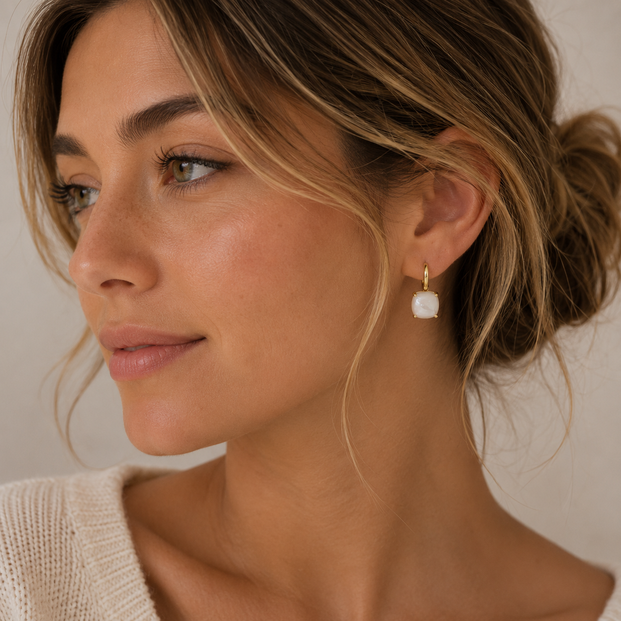 A woman with light brown hair in a loose bun, wearing a cream sweater and JaxKelly’s Gemstone Mini Drop - Mother of Pearl earrings, gazes thoughtfully to the side against a soft beige background.