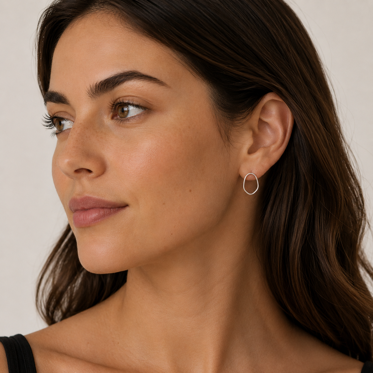 A woman with long brown hair gazes to the side, wearing a black top and the JaxKelly Silver Organic Hoop earrings. The background is neutral and softly lit.