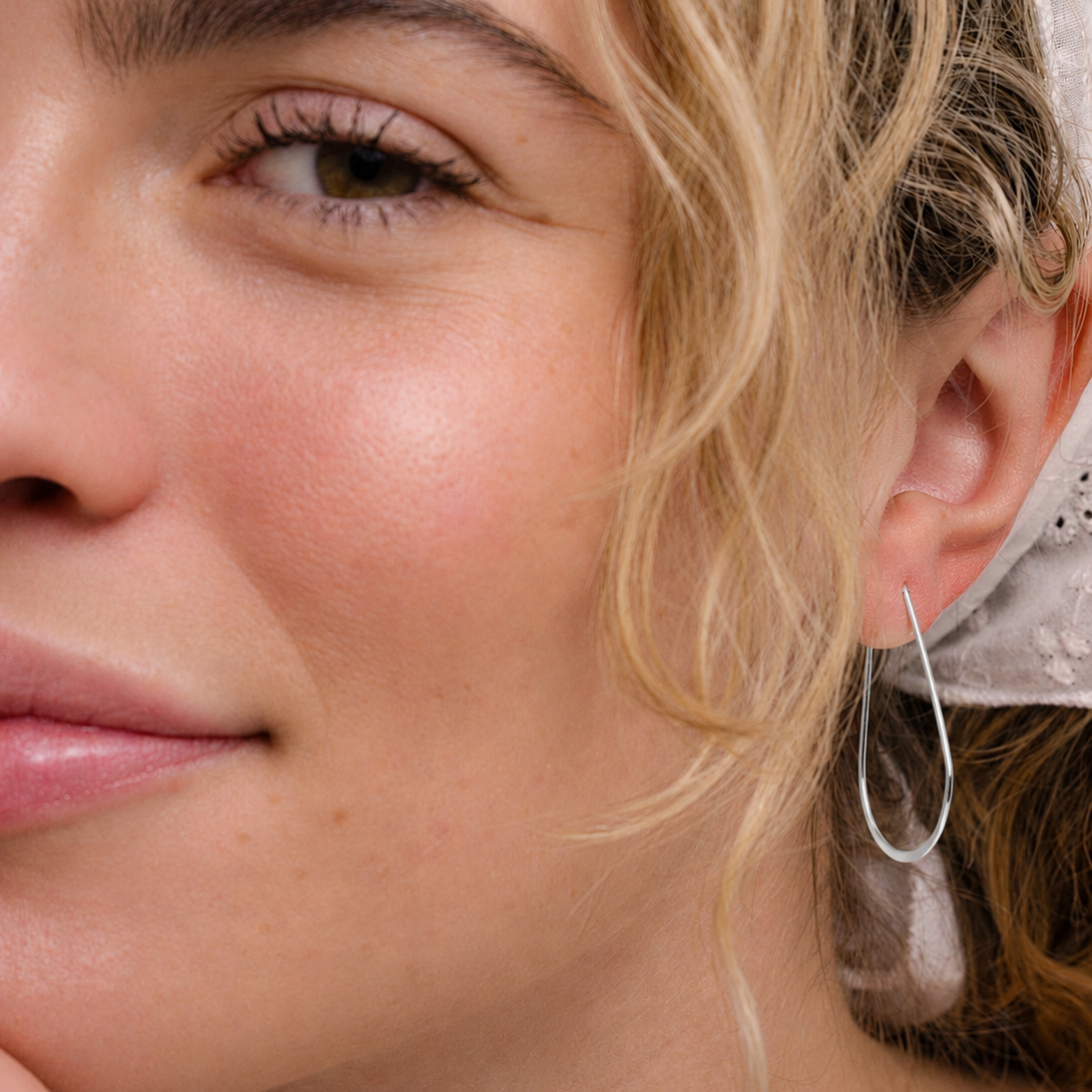 A close-up of a woman with light skin, blonde wavy hair, and a white scarf, smiling softly while wearing JaxKelly's Silver Long Oval Hoop earrings.