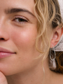 A close-up of a woman with light skin, blonde wavy hair, and a white scarf, smiling softly while wearing JaxKelly's Silver Long Oval Hoop earrings.