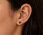 Close-up of a person with straight dark hair wearing the JaxKelly Black Baguette stud earring—a small rectangular black gemstone set in gold with sterling silver posts. The background is blurred.