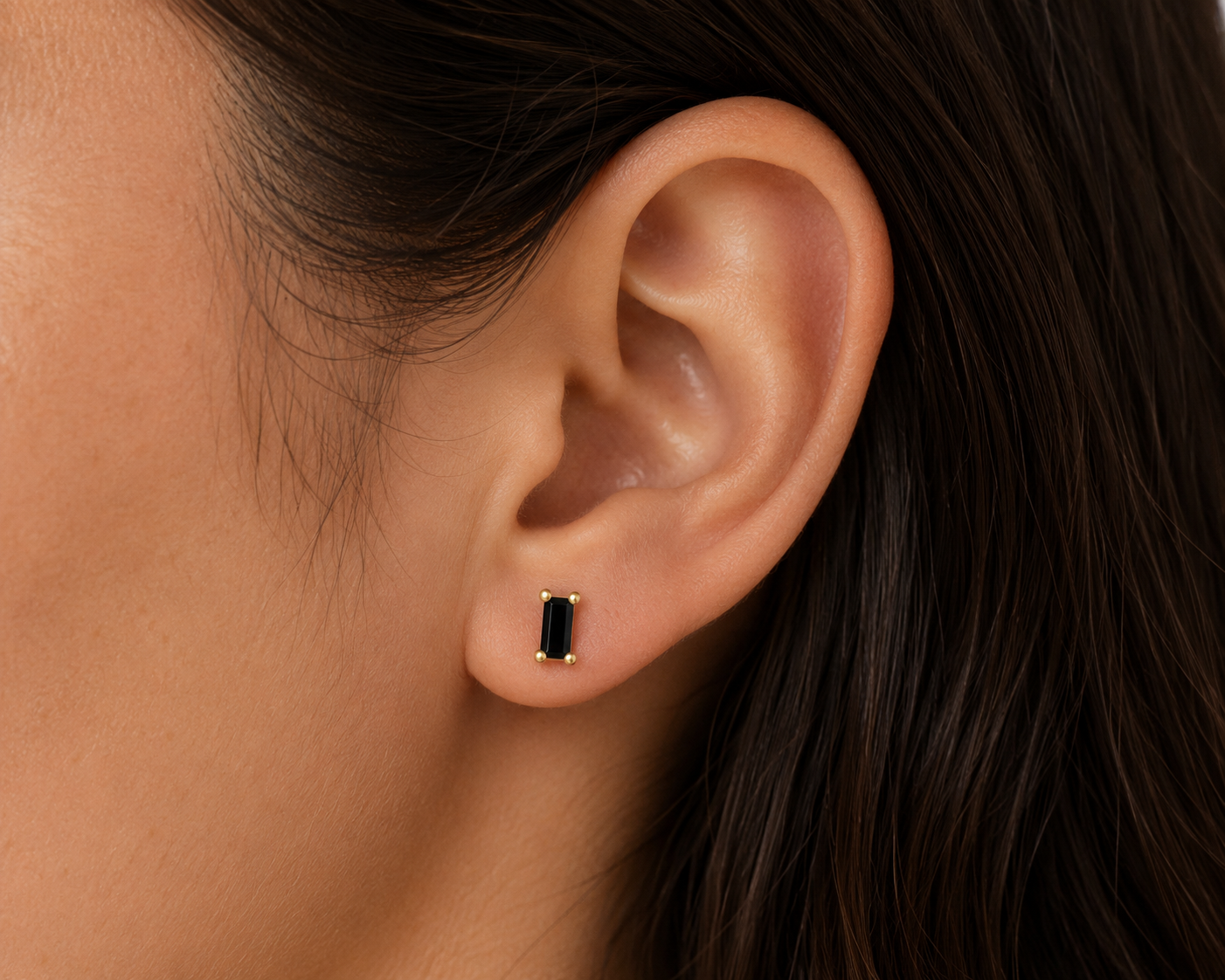 Close-up of a person with straight dark hair wearing the JaxKelly Black Baguette stud earring—a small rectangular black gemstone set in gold with sterling silver posts. The background is blurred.