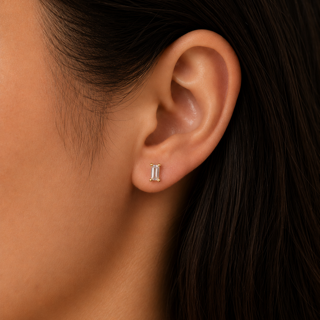 Close-up of a woman's ear with straight dark hair, wearing JaxKelly's Clear Baguette stud earring. This small, hypoallergenic design offers a stylish and comfortable look with its minimalist clear baguette detailing.
