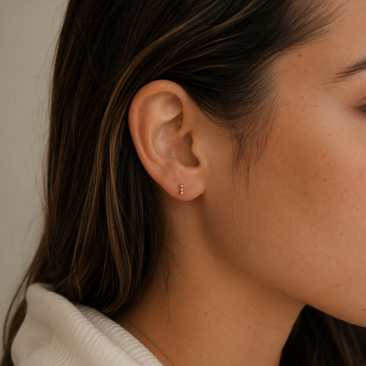 A close-up of a woman's ear with long brown hair wearing the JaxKelly Petite 3 Ball Stud, a minimalist gold-plated, hypoallergenic earring, against a neutral background. Part of her face and shoulder are also visible.