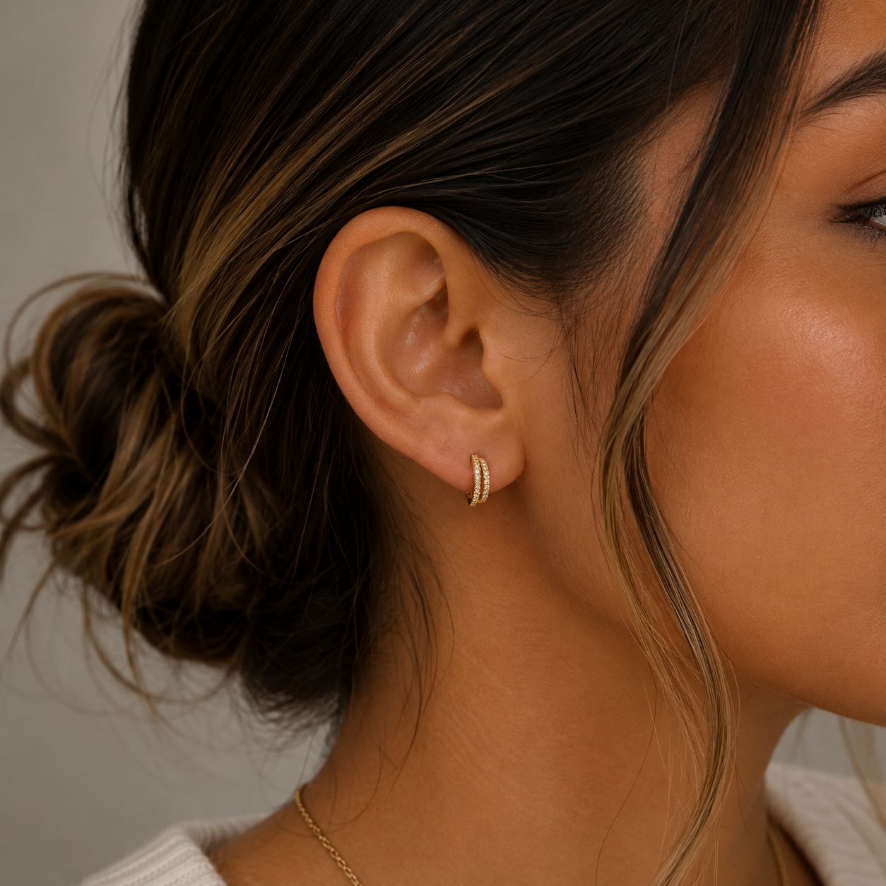 A close-up of a woman wearing JaxKelly's Double Champagne Hoop Earrings. The 18K gold-plated hoops shine as her dark hair is styled in a loose bun, paired with a white top and gold necklace against a neutral background.