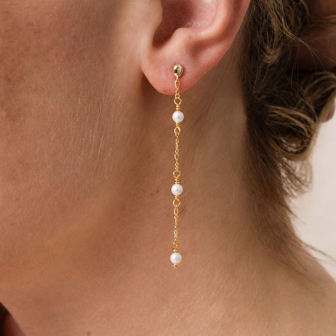 Close-up of a woman wearing JaxKelly's Gemstone Chain Drop - Mother of Pearl earring, featuring three white pearls on a delicate gemstone chain and finished with 18k gold plating, gracefully hanging past her earlobe. Part of her face and hair are visible.