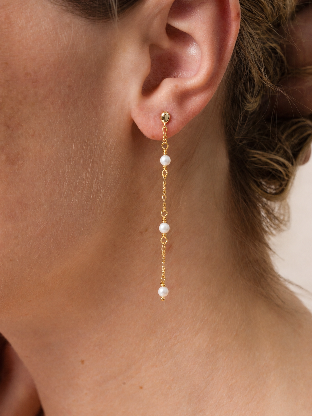 Close-up of a woman wearing JaxKelly's Gemstone Chain Drop - Mother of Pearl earring, featuring three white pearls on a delicate gemstone chain and finished with 18k gold plating, gracefully hanging past her earlobe. Part of her face and hair are visible.