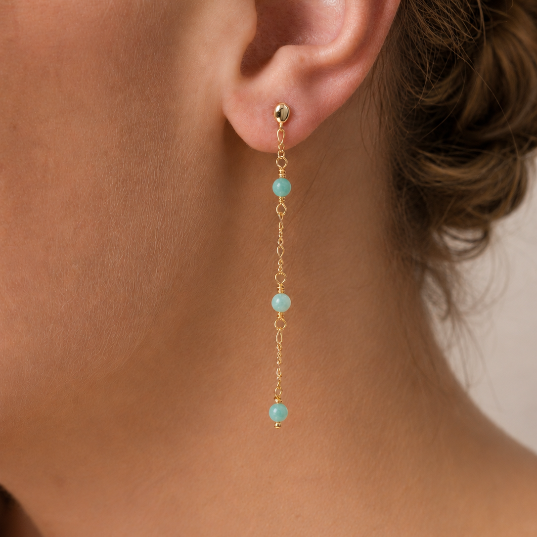 Close-up of a woman's ear wearing JaxKelly's Gemstone Chain Drop - Amazonite: a long 18k gold plated earring with three Amazonite beads spaced along a delicate chain. Her hair is pulled back against a neutral background.