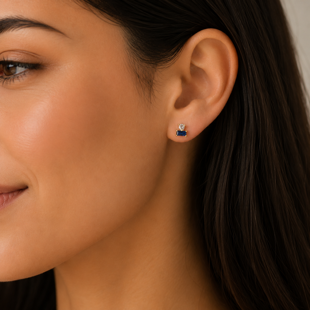 A close-up of a woman with smooth skin and straight dark brown hair wearing the JaxKelly Sapphire Double Stack stud, featuring a rectangular sapphire set in 18k gold plating with a small round CZ stone above it.