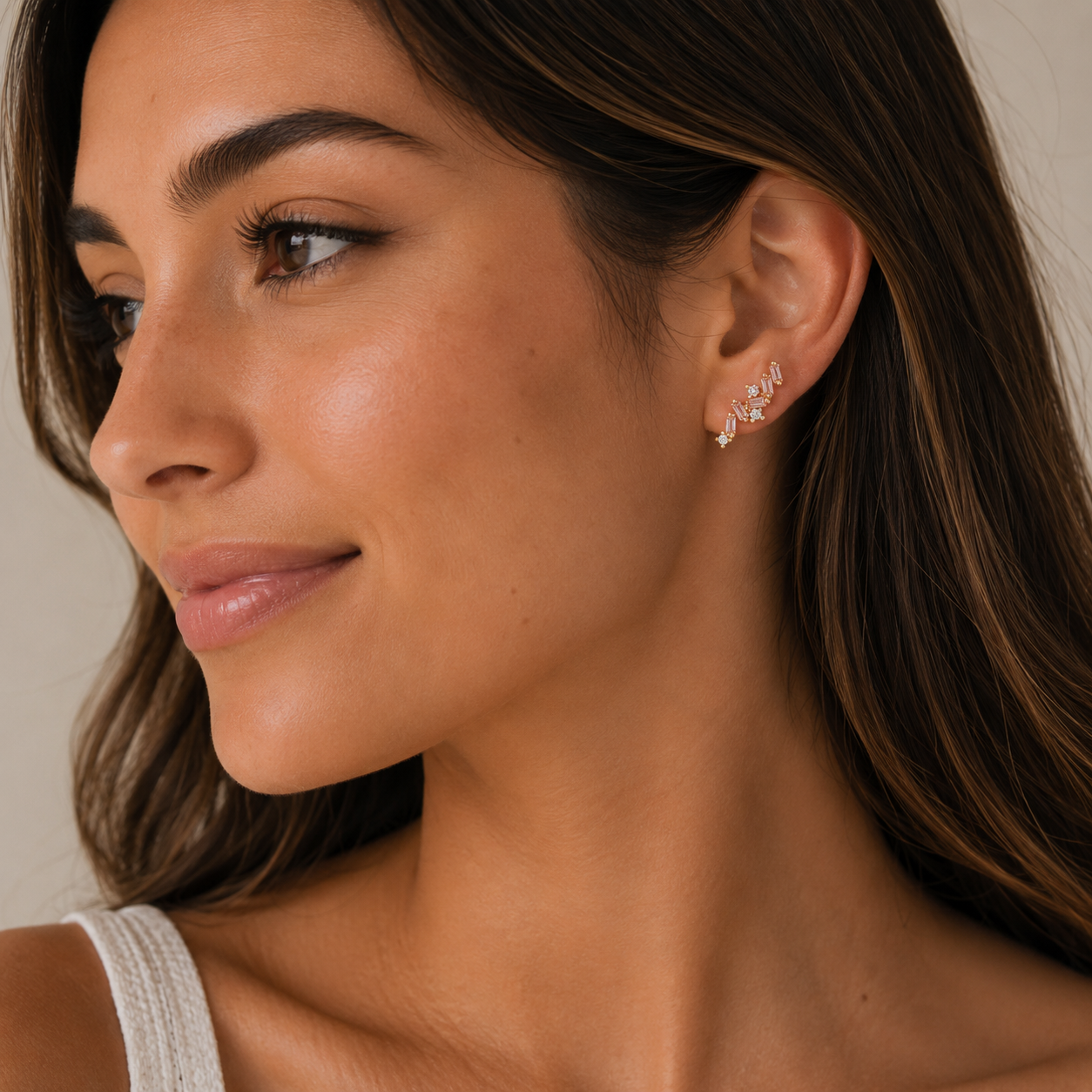 A woman with long brown hair gazes to the side, wearing a sleeveless top and JaxKelly's Cascading Baguette Drop - Gold Earrings that shimmer delicately. Her natural makeup and subtle smile complete her elegant look.
