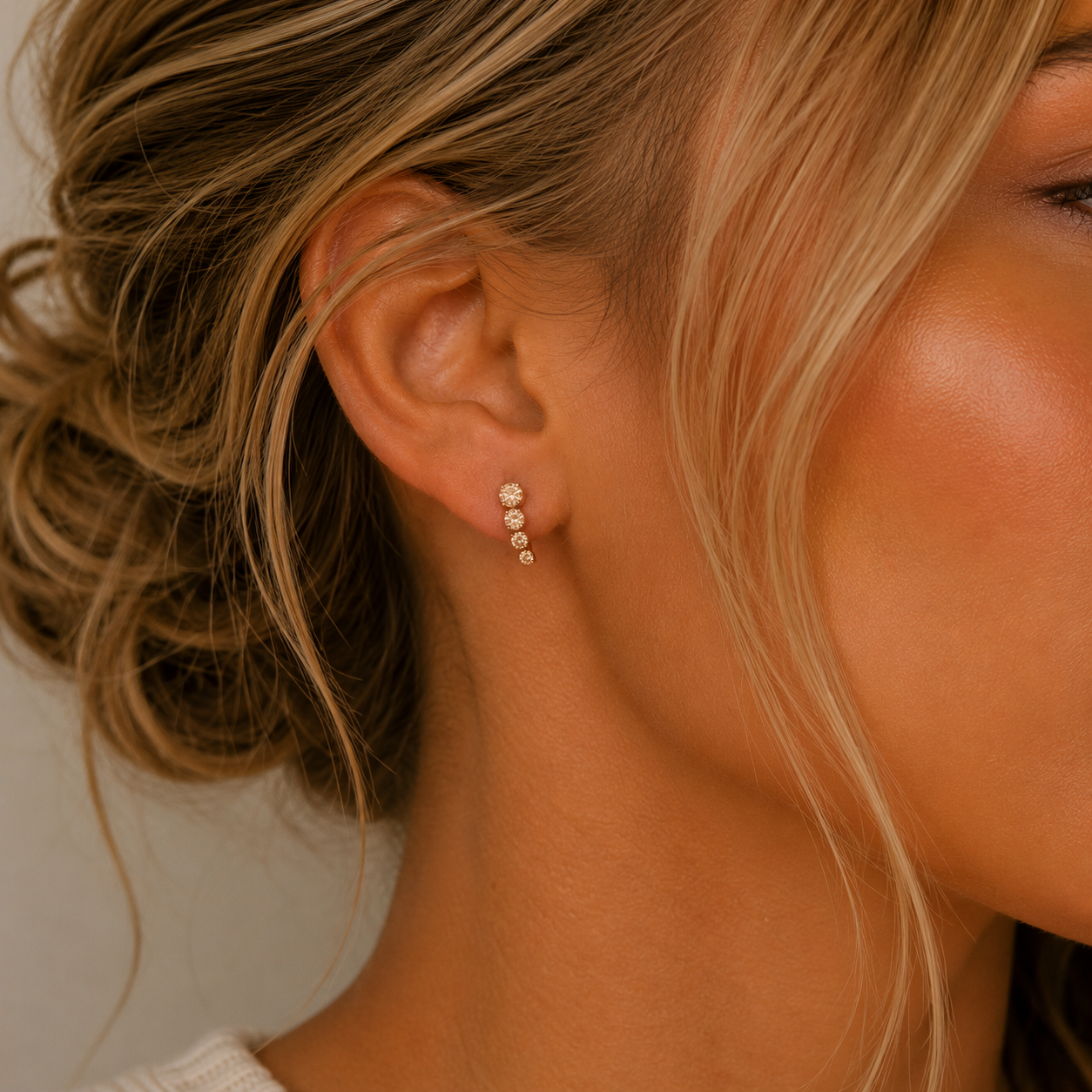 Close-up of a woman's ear with blonde hair in a loose updo, wearing JaxKelly's Mini Crawler - Champagne CZ - Gold Earrings. Three sparkling champagne CZ stones are arranged vertically. The background is neutral and softly blurred.