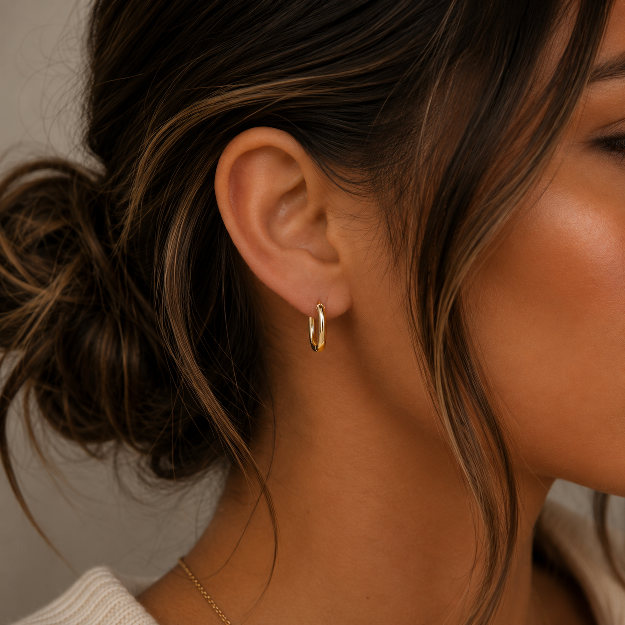 A close-up of a woman's ear shows her wearing the JaxKelly Color Dipped Rectangle Hoop Earring in brown. Her brown hair is styled in a messy bun against a neutral, blurred background, highlighting the unique design.