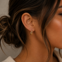A close-up of a woman's ear shows her wearing the JaxKelly Color Dipped Rectangle Hoop Earring in brown. Her brown hair is styled in a messy bun against a neutral, blurred background, highlighting the unique design.