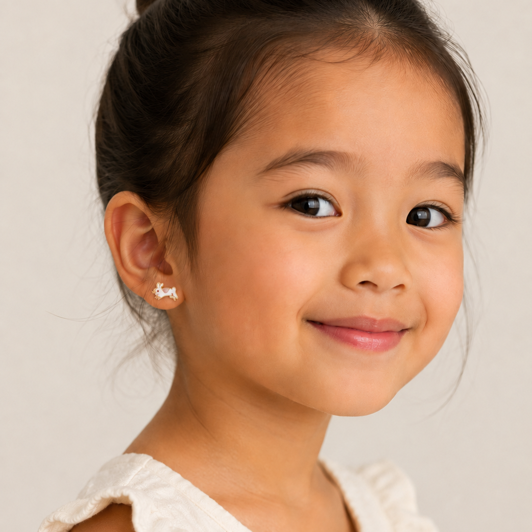 A young girl with dark hair in a bun smiles gently, wearing a cream top with ruffled sleeves and JaxKelly Bunny Enamel Flat Back Kids Earrings—hypoallergenic and perfect for sensitive ears—against a light neutral background.