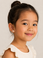 A young girl with dark hair in a bun smiles gently, wearing a cream top with ruffled sleeves and JaxKelly Bunny Enamel Flat Back Kids Earrings—hypoallergenic and perfect for sensitive ears—against a light neutral background.