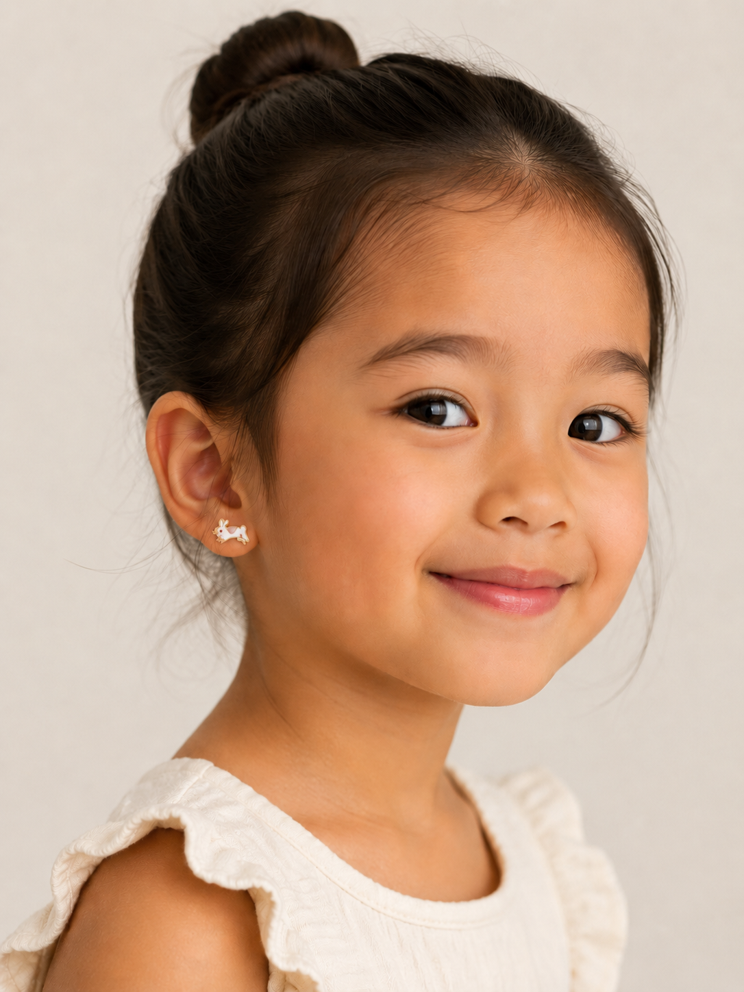 A young girl with dark hair in a bun smiles gently, wearing a cream top with ruffled sleeves and JaxKelly Bunny Enamel Flat Back Kids Earrings—hypoallergenic and perfect for sensitive ears—against a light neutral background.