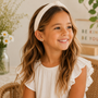 A young girl with long wavy hair and a cream headband smiles in a wicker chair, wearing a white ruffled dress and JaxKelly Rainbow Enamel Flat Back Kids Earrings. Daisies in a vase and a “Be Kind” sign add charm to the scene.