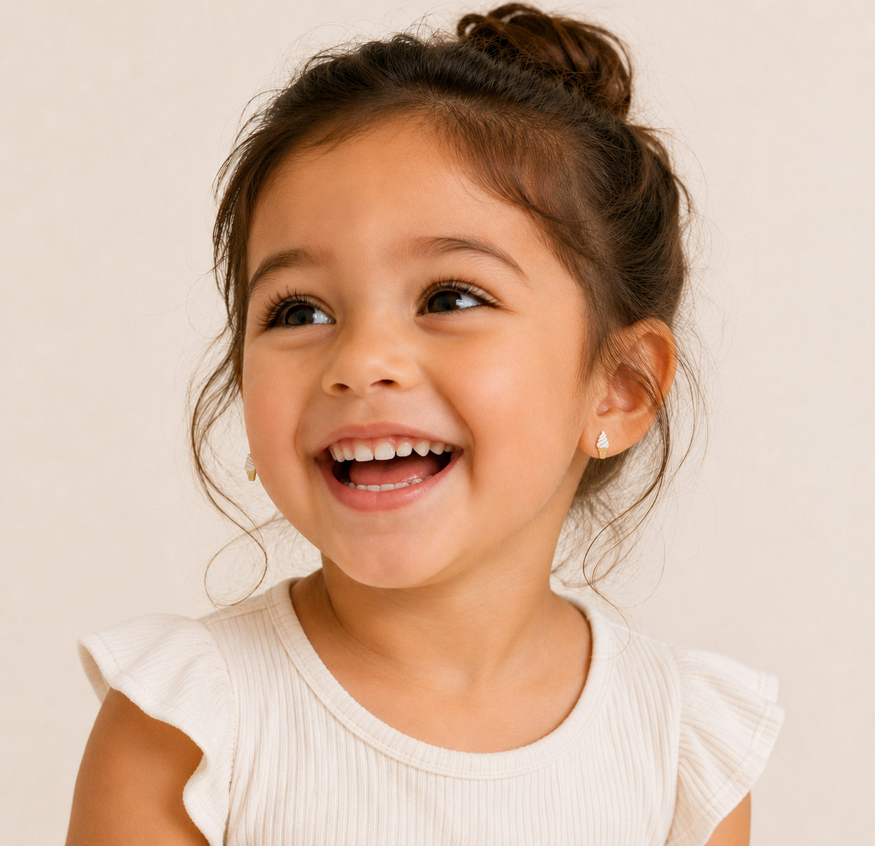 A young girl with dark hair in a messy bun and a white top smiles brightly, showcasing JaxKelly's Ice Cream Enamel Flat Back Kids Earrings—hypoallergenic and perfect for sensitive ears—while looking slightly to the side against a neutral background.