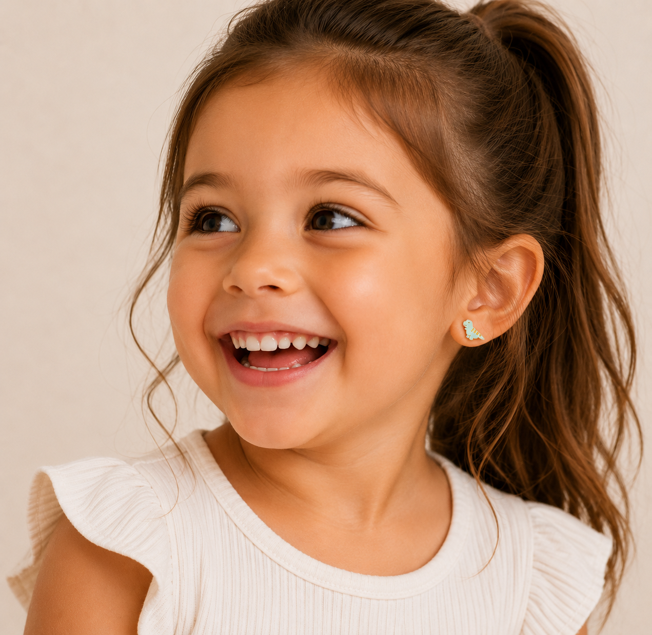 A smiling young girl with long brown hair in a ponytail wears a sleeveless cream ruffled top and JaxKelly Dinosaur Enamel Flat Back Kids Earrings. The background is plain and neutral.
