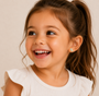 A smiling young girl with long brown hair in a ponytail wears a sleeveless cream ruffled top and JaxKelly Dinosaur Enamel Flat Back Kids Earrings. The background is plain and neutral.