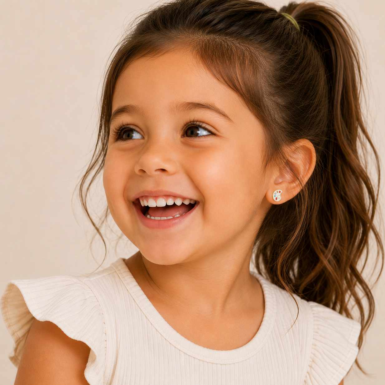 A smiling young girl with long brown hair in a ponytail, wearing a cream top with ruffled sleeves, looks to the side. She wears JaxKelly Artist Palette Enamel Flat Back Kids Earrings, designed for sensitive ears.