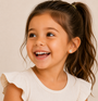 A smiling young girl with long brown hair in a ponytail, wearing a cream top with ruffled sleeves, looks to the side. She wears JaxKelly Artist Palette Enamel Flat Back Kids Earrings, designed for sensitive ears.