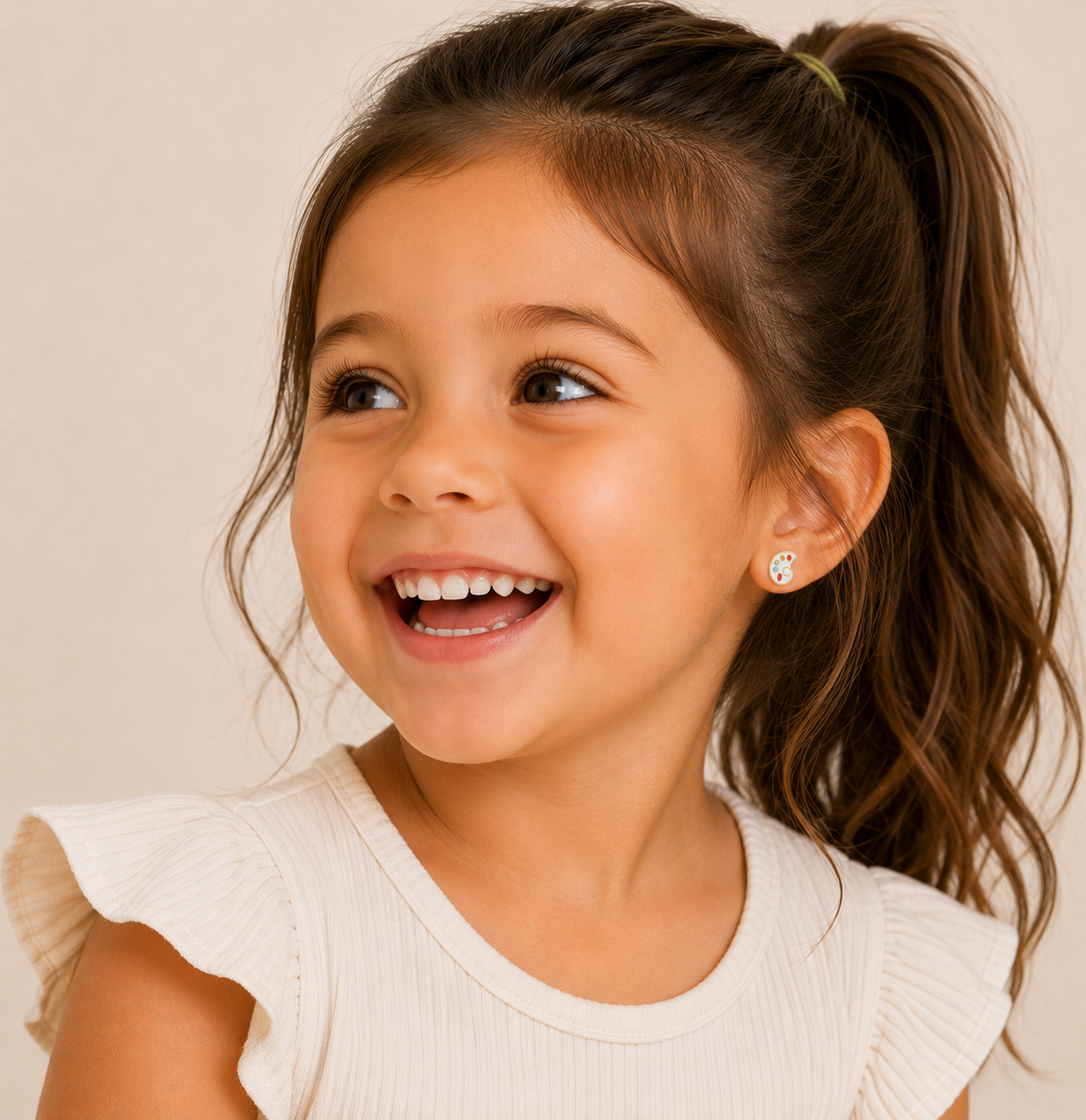 A smiling young girl with long brown hair in a ponytail, wearing a cream top with ruffled sleeves, looks to the side. She wears JaxKelly Artist Palette Enamel Flat Back Kids Earrings, designed for sensitive ears.