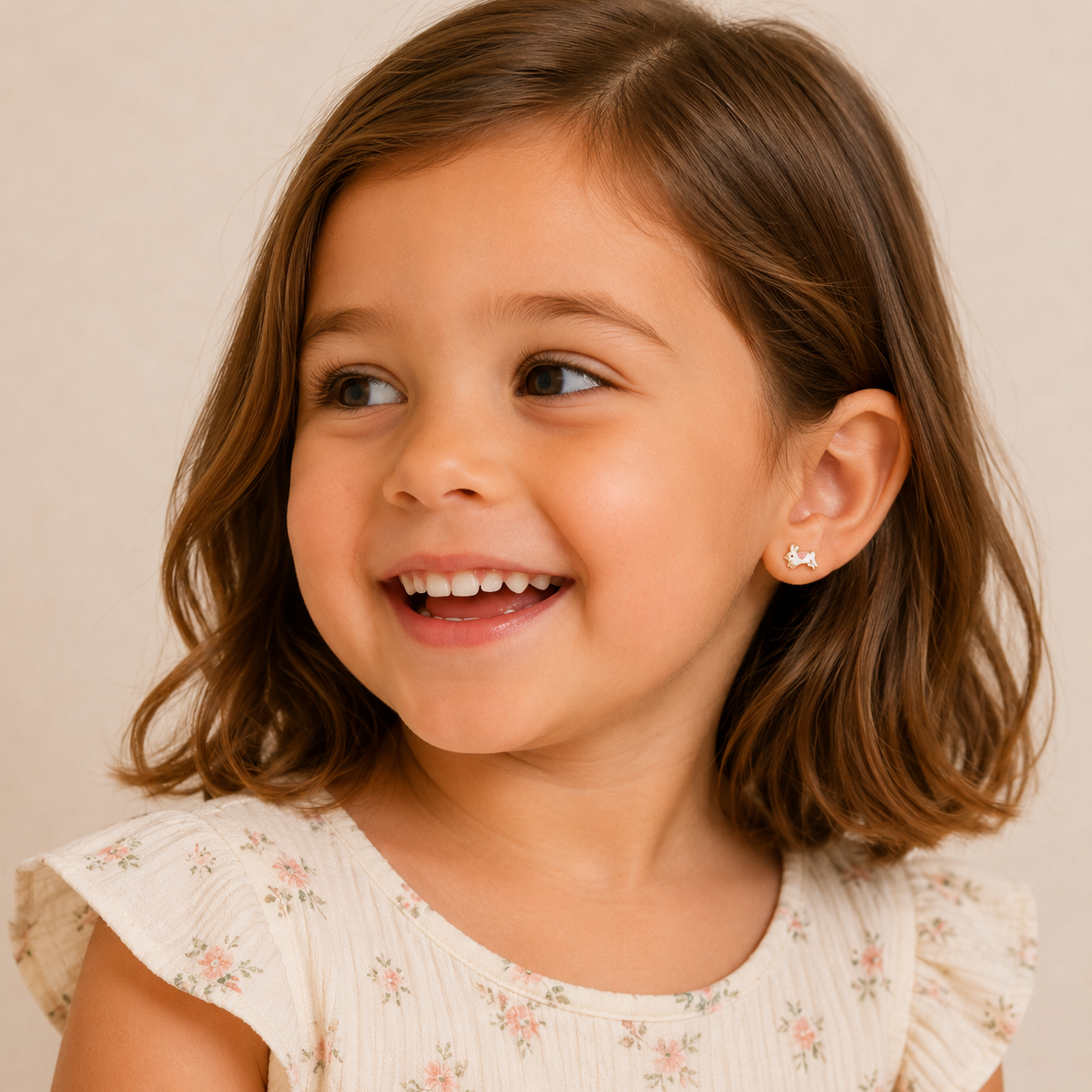 A young girl with brown bobbed hair smiles while looking to the side. She wears a light floral dress and JaxKelly Bunny Enamel Flat Back Kids Earrings, made from surgical-grade stainless steel. The softly lit background is neutral.