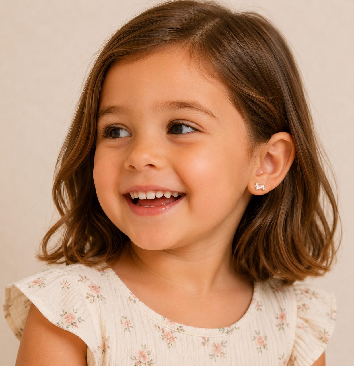A young girl with brown bobbed hair smiles while looking to the side. She wears a light floral dress and JaxKelly Bunny Enamel Flat Back Kids Earrings, made from surgical-grade stainless steel. The softly lit background is neutral.