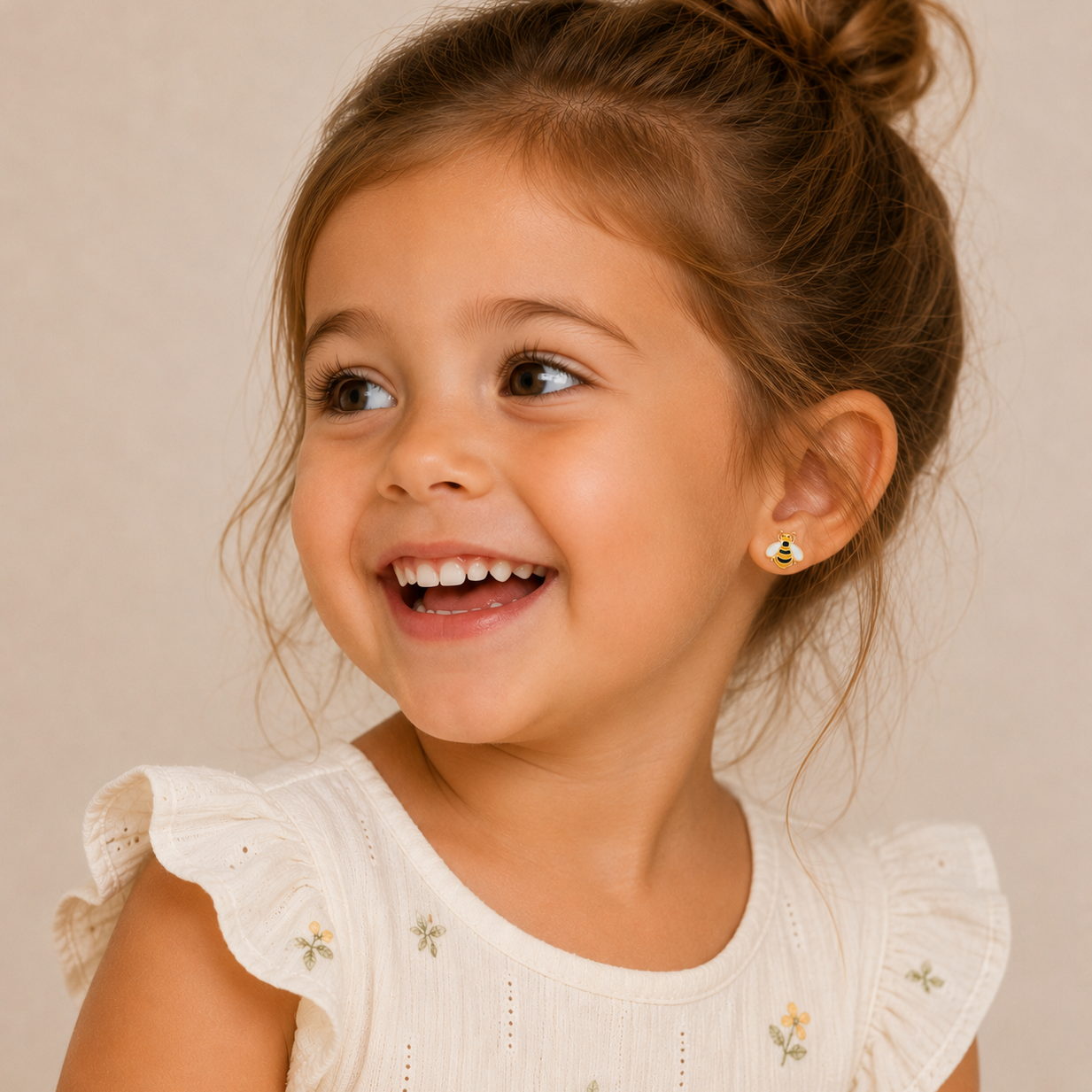 A young girl with brown hair in a bun smiles happily, showing off her JaxKelly Bumble Bee Enamel Flat Back Kids Earrings—perfect hypoallergenic earrings for sensitive ears. She wears a cream dress with ruffled sleeves and floral embroidery.