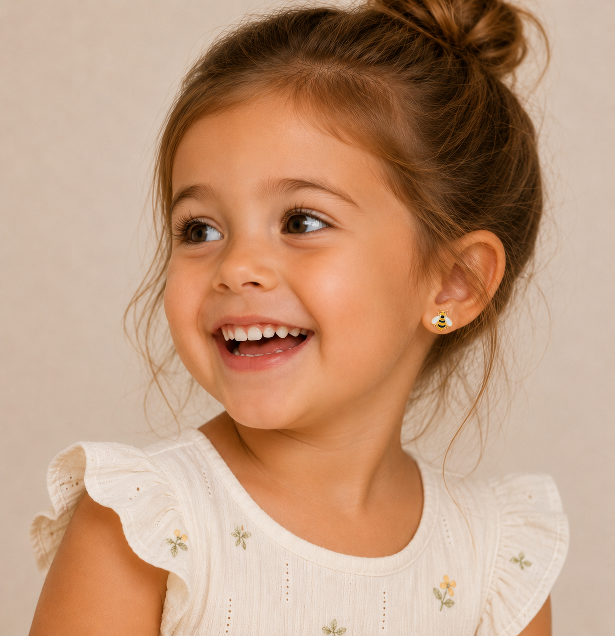 A young girl with brown hair in a bun smiles happily, showing off her JaxKelly Bumble Bee Enamel Flat Back Kids Earrings—perfect hypoallergenic earrings for sensitive ears. She wears a cream dress with ruffled sleeves and floral embroidery.