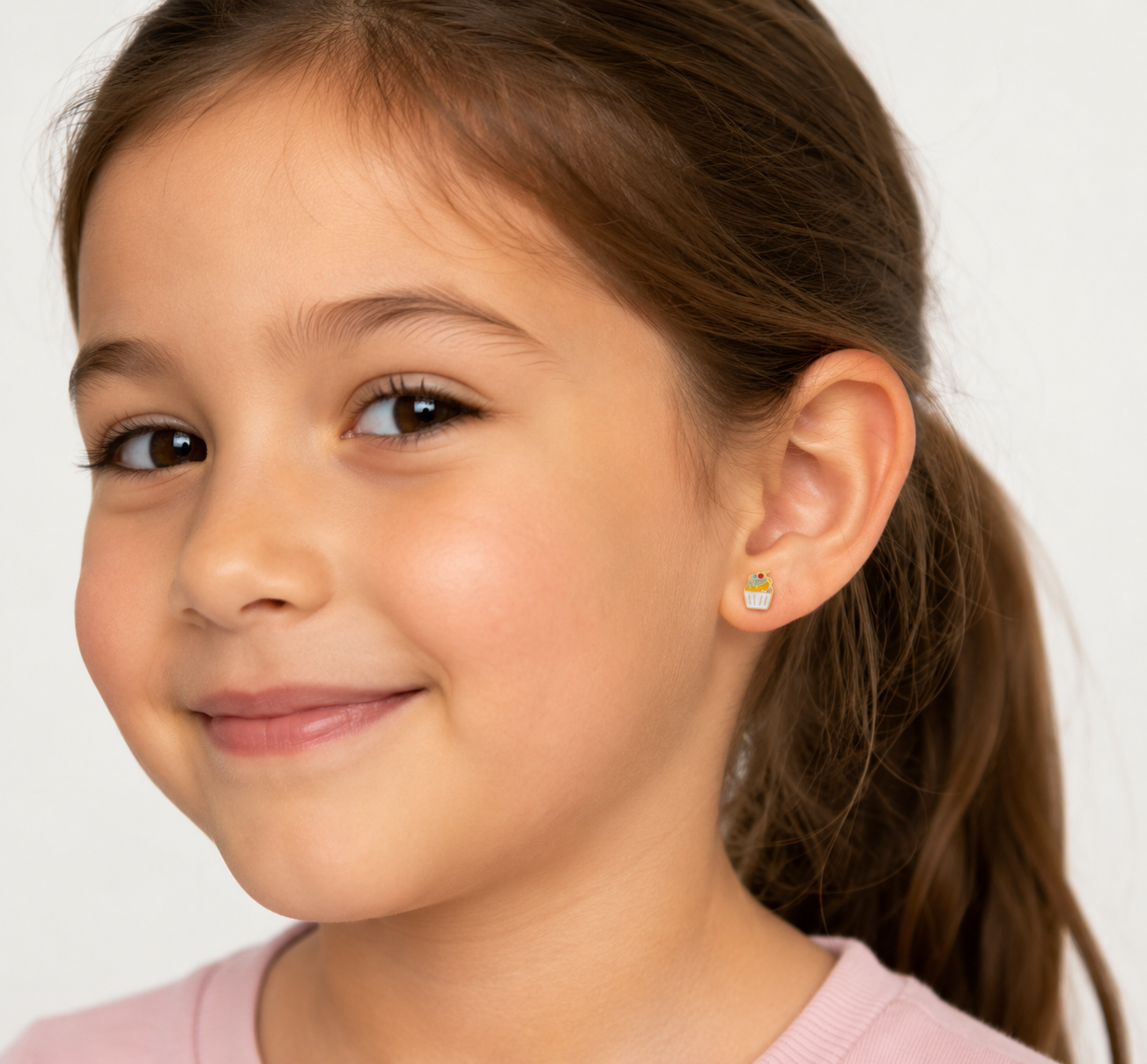 A young girl with long brown hair smiles gently, wearing JaxKelly’s Cupcake Enamel Flat Back Kids Earrings for sensitive ears. She is shown from the shoulders up against a plain light background.