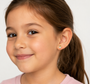 A young girl with long brown hair smiles gently, wearing JaxKelly’s Cupcake Enamel Flat Back Kids Earrings for sensitive ears. She is shown from the shoulders up against a plain light background.