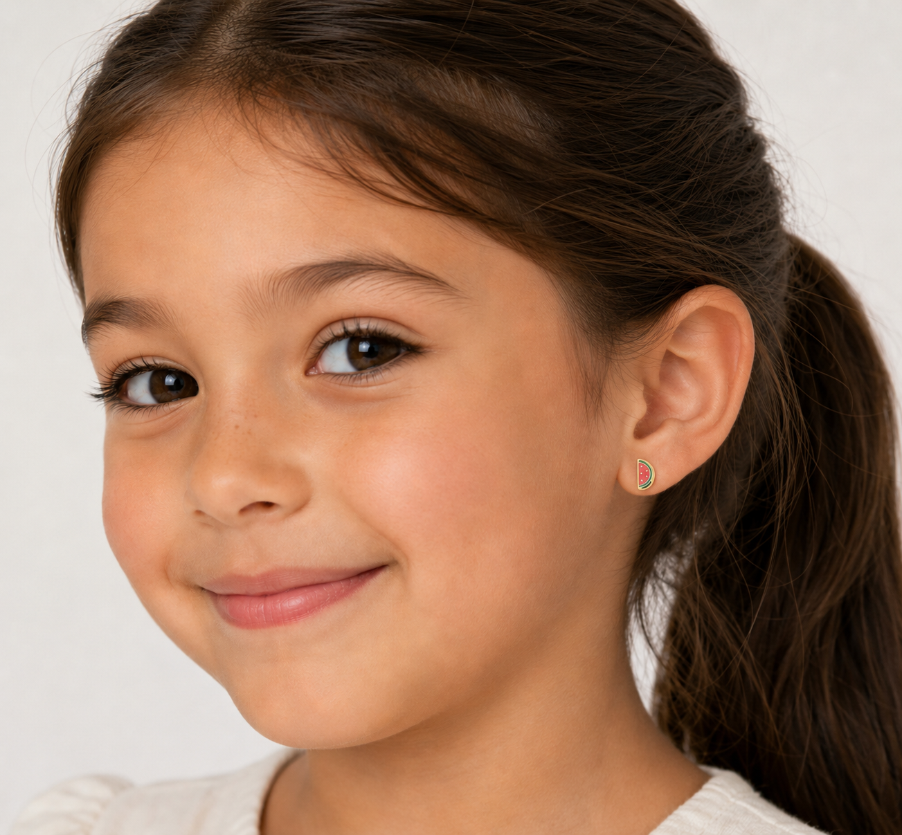 A young girl with long brown hair smiles softly, wearing a cream top and JaxKelly’s Watermelon Enamel Flat Back Kids Earring—perfect for sensitive ears. The background is plain and light-colored.