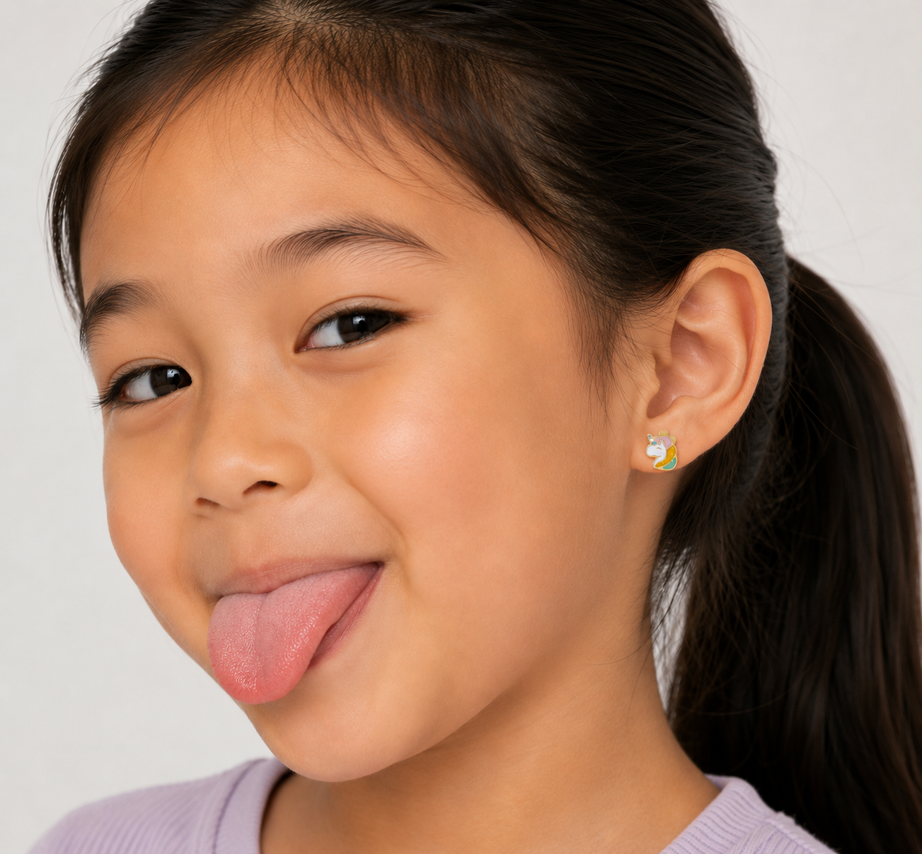 A young girl with long dark hair in a ponytail smiles playfully, wearing a lavender top and JaxKelly Unicorn Enamel Flat Back Kids Earrings—hypoallergenic and perfect for sensitive ears. The plain background is light-colored.