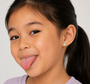 A young girl with long dark hair in a ponytail smiles playfully, wearing a lavender top and JaxKelly Unicorn Enamel Flat Back Kids Earrings—hypoallergenic and perfect for sensitive ears. The plain background is light-colored.