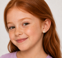 A young girl with long red hair and freckles smiles softly, wearing a lavender shirt and JaxKelly Heart Enamel Flat Back Kids Earrings, perfect for sensitive ears, as she faces slightly to the side against a neutral background.