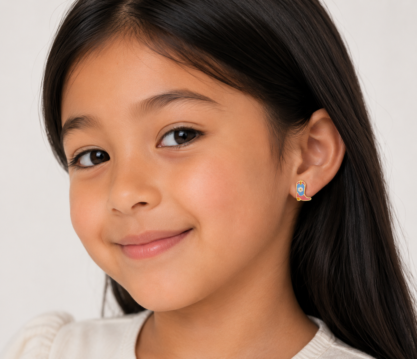 A young girl with long dark hair and a slight smile wears cream and Boot Enamel Flat Back Kids Earrings by JaxKelly—colorful bird-shaped studs perfect for sensitive ears. The background is plain and light.